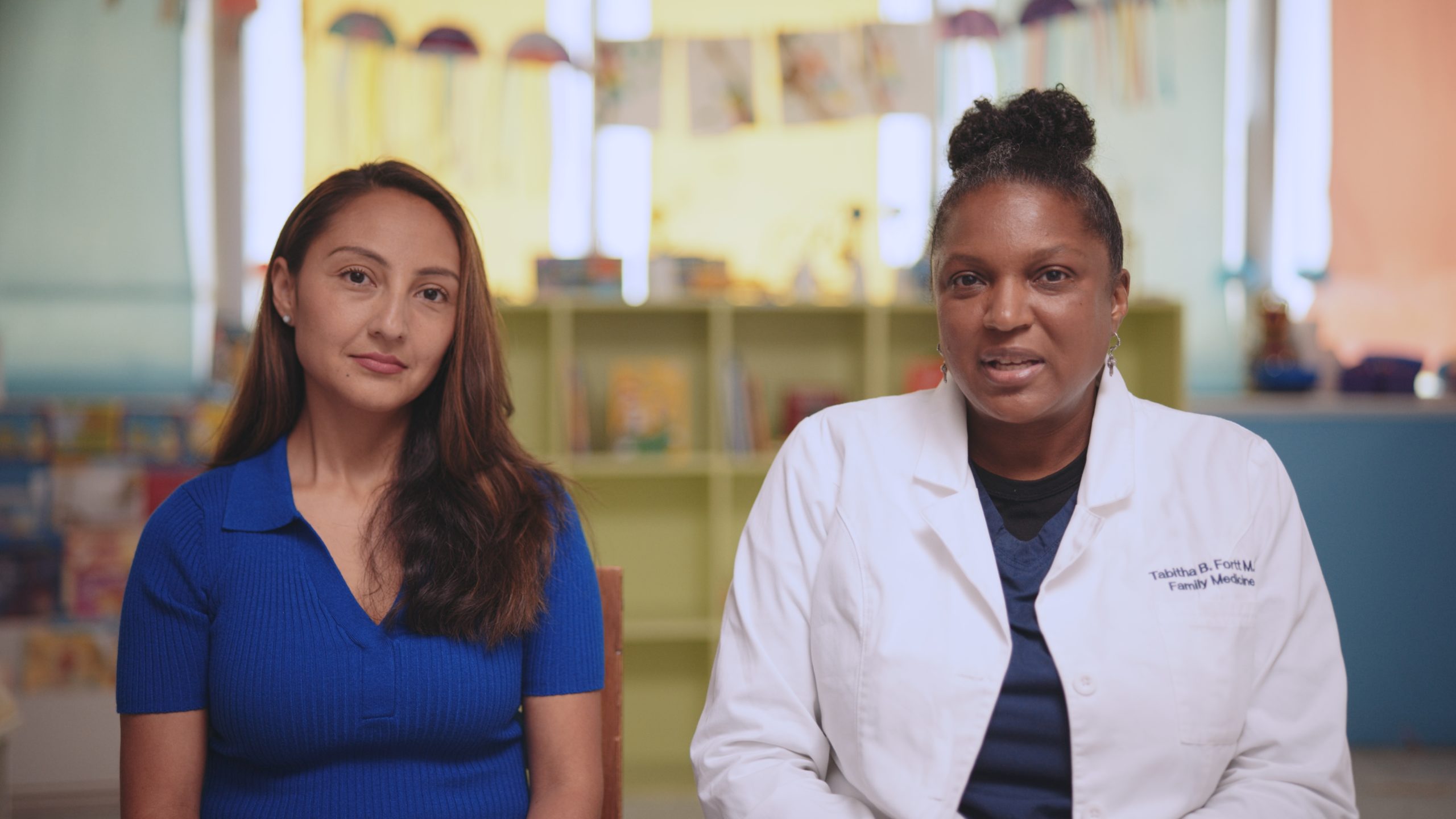 IU C&I Studios Page Conceptual Communications Woman wearing blue top with long brown hair sitting next to African American female family medicine doctor with a hair in a bun wearing a white coat posing for the camera