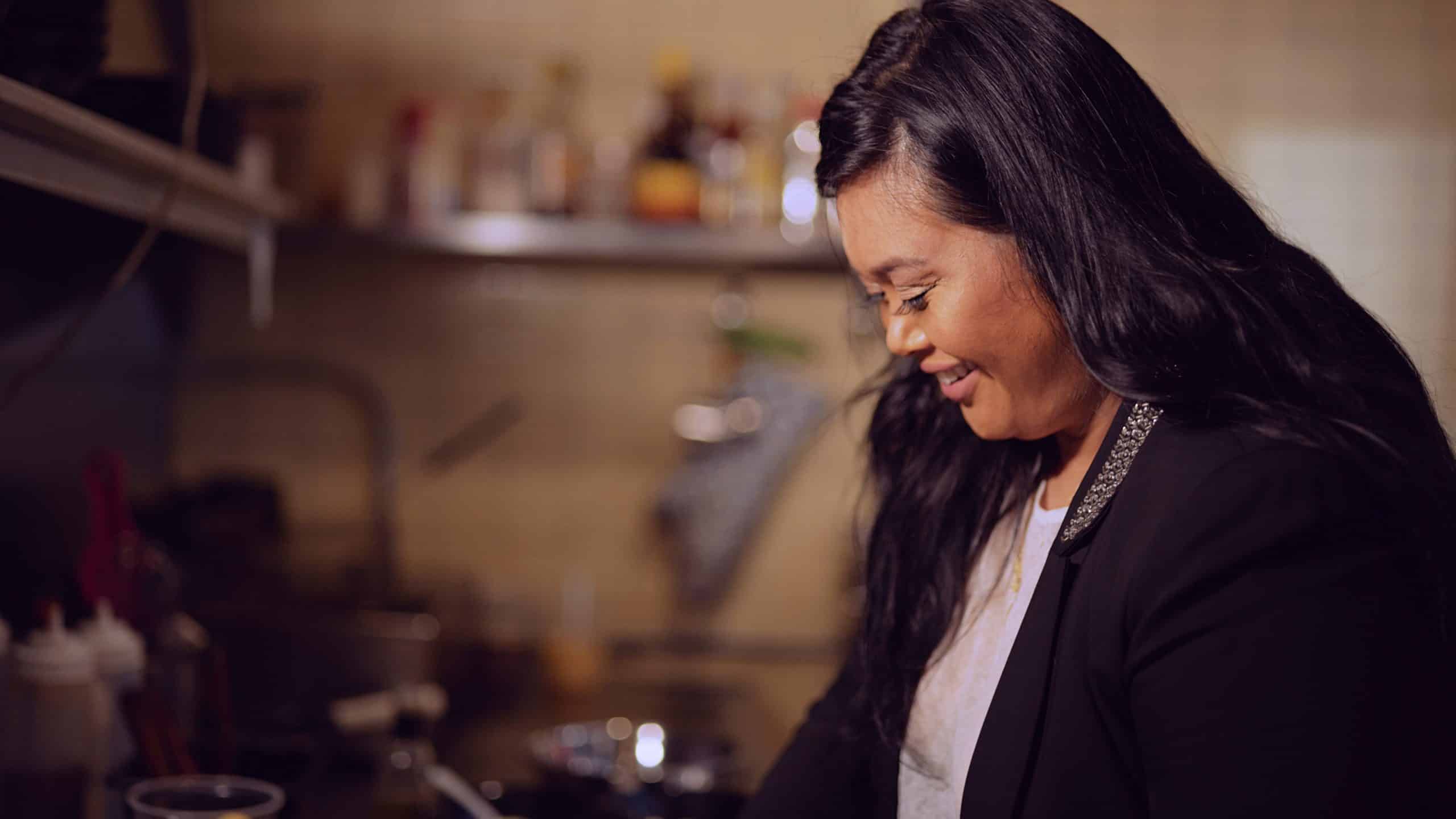 Eye-Catching Jeepney NYC Filipino BBQ Pop Up Restaurant Side profile headshot of female chef with long black hair working in the kitchen of a restaurant smiling