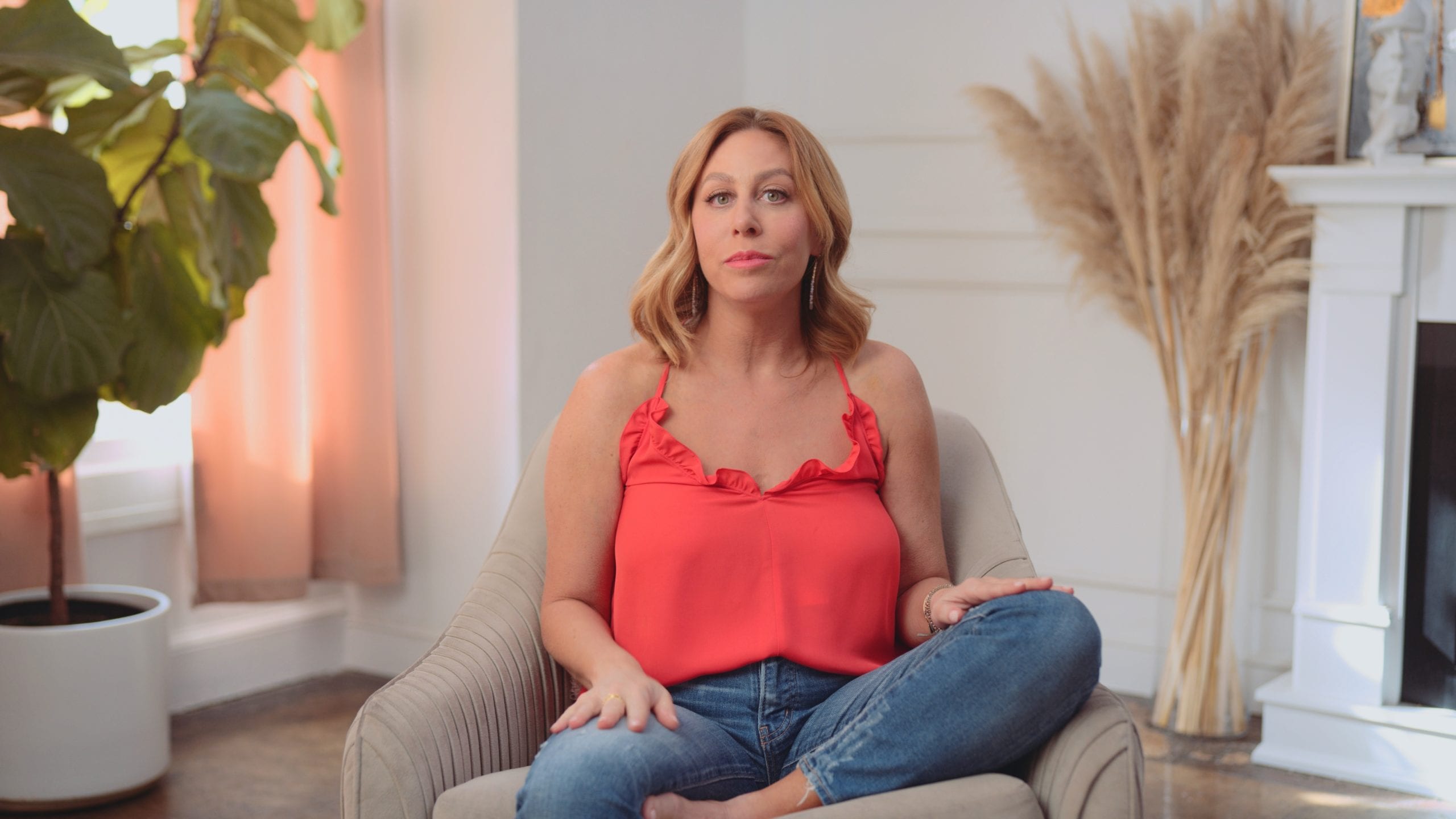 Woman with long blond hair wearing an orange top and jeans talking to camera sitting on a beige chair