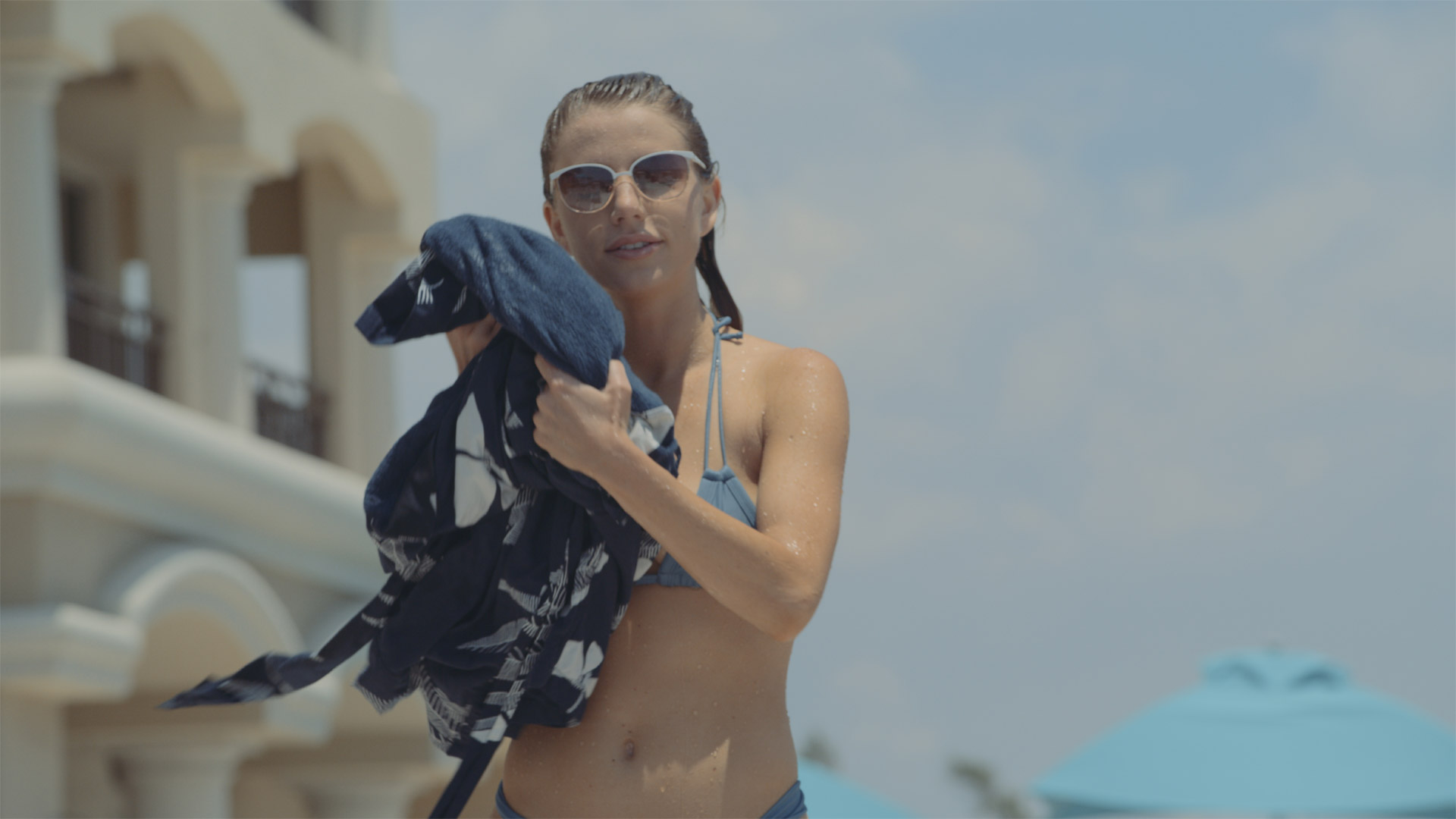 Woman wearing blue bikini and sunglasses posing for camera holding a beach outfit in her hands