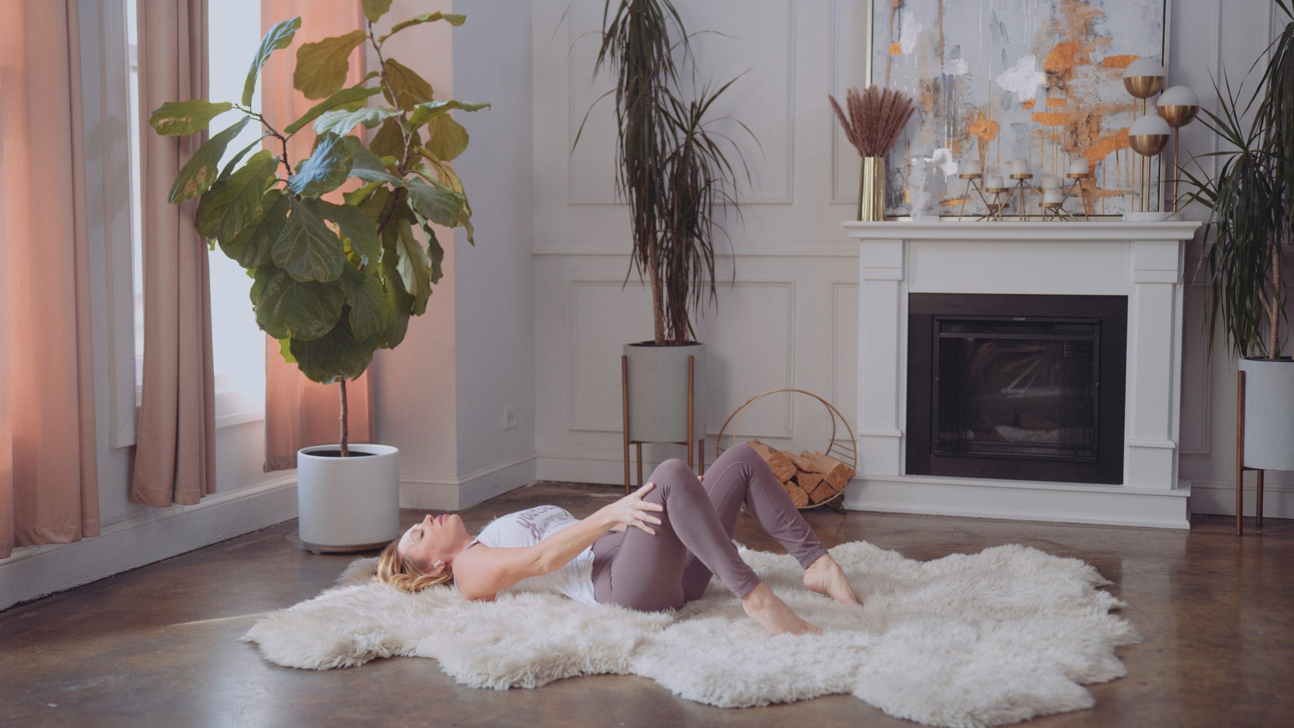 Woman with long blond hair wearing white top and purple tights laying on a white fluffy rug with eyes closed and knees up