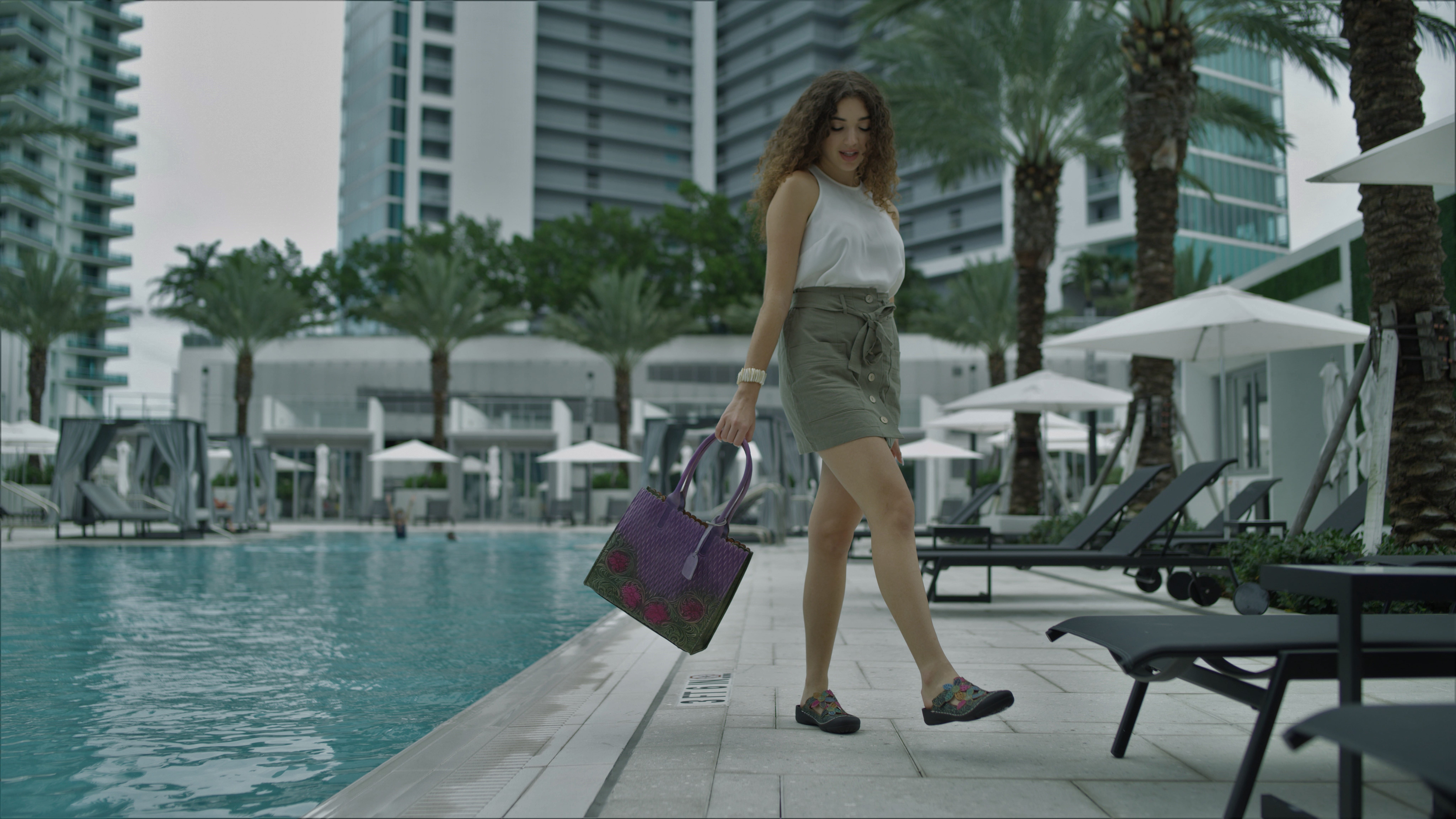 Spring StepLartiste Kalria Side profile of woman wearing white dress top, olive green shorts carrying a purple purse walking by a pool with black lounge chairs and patio umbrellas nearby