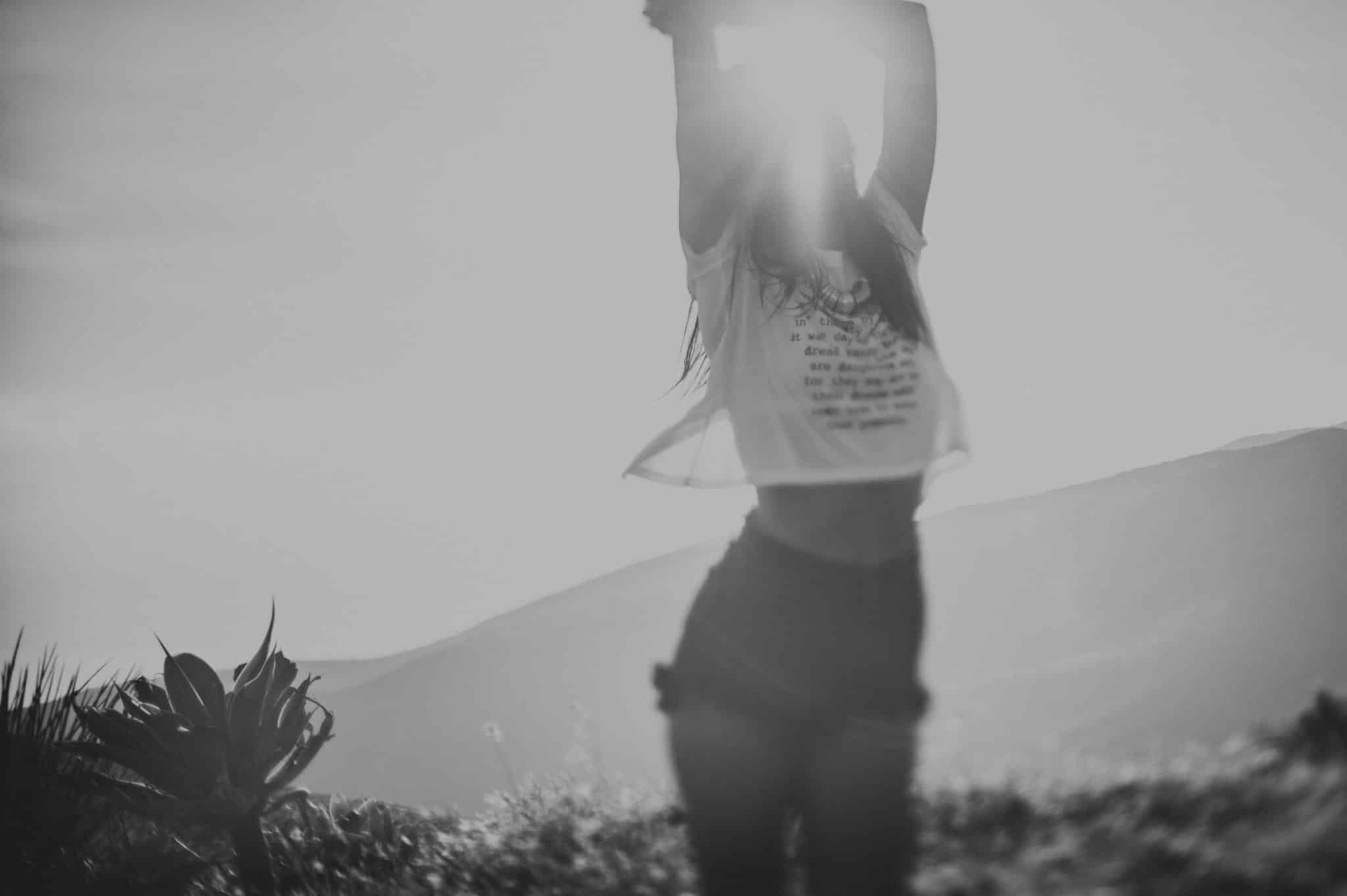 Blurry black and white photo of a young female wearing a short white top with writing and short black shorts. She has her arms raised in the air with the sun filtering through.