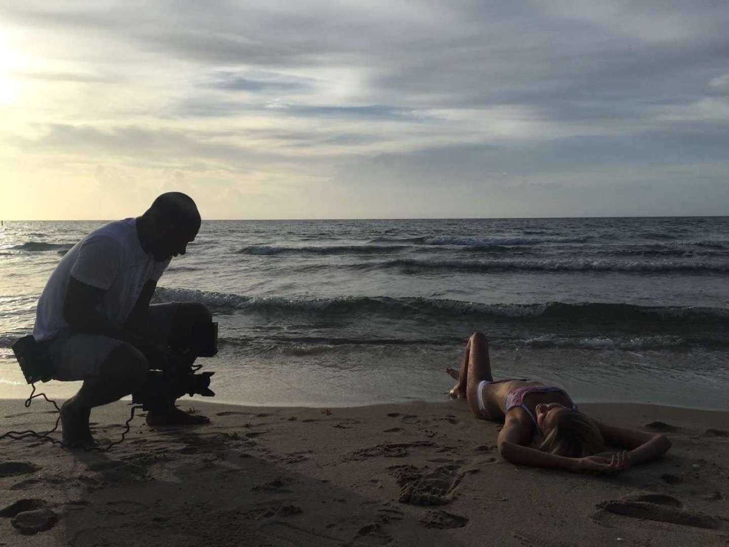 CI Studios Portfolio Taylor Adair Swim BTS Young woman with long blond hair in a bikini laying on the sand at a beach near the water posing for the camera man with her arms behind her head