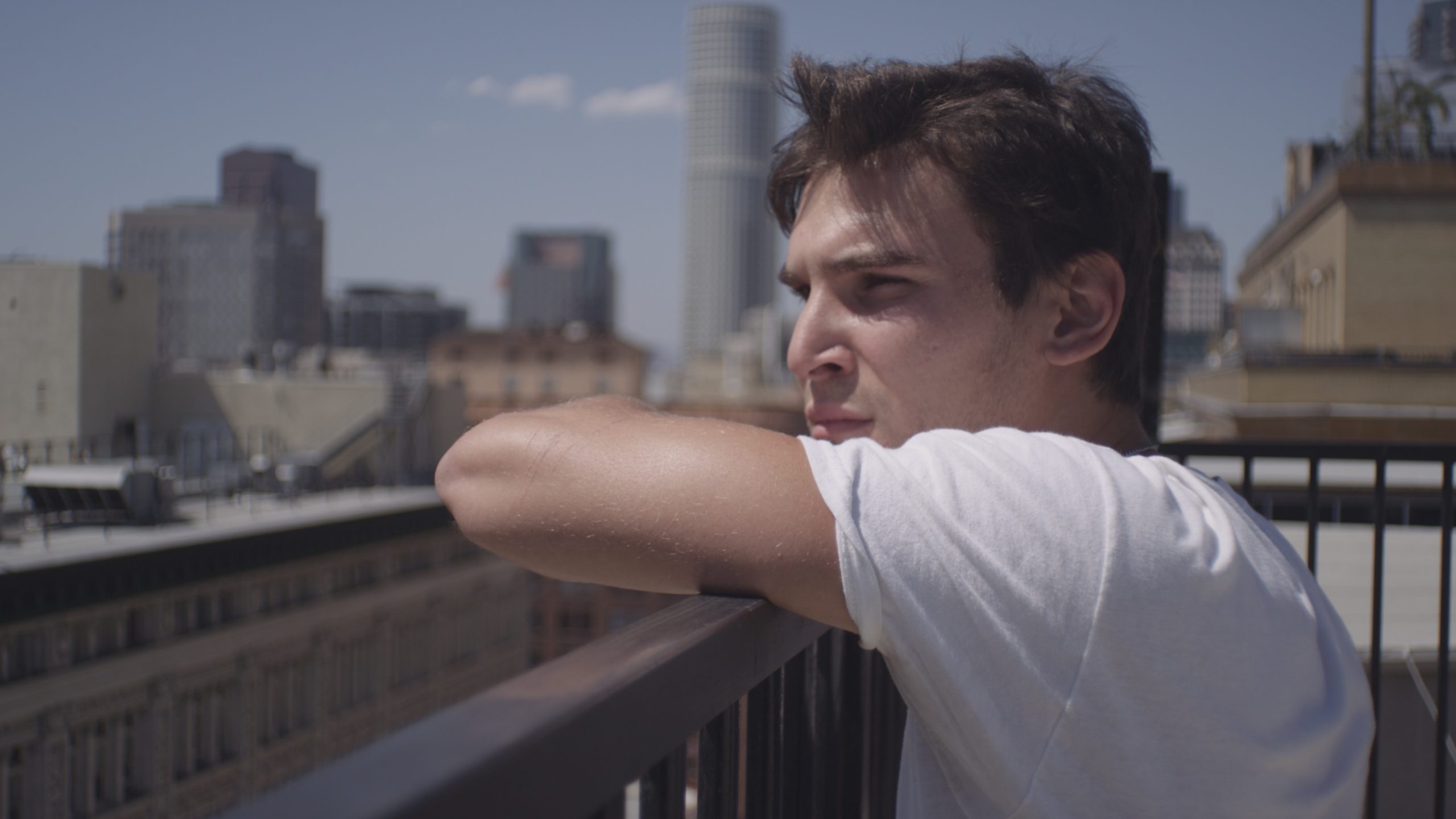 Uncreative Stories Joe Perri Profile headshot of man with short brown hair wearing a white t shirt looking out over a city leaning on a metal railing