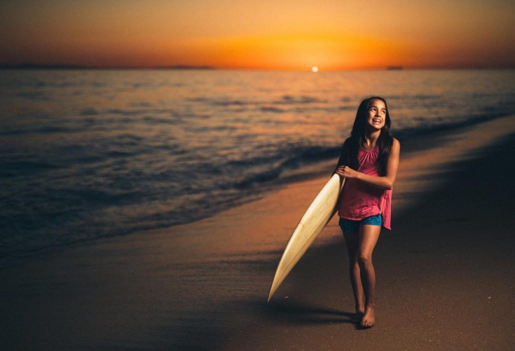 Ivivva Lulu Lemon Girl on a beach carrying a surfboard at dusk looking off to the side and smiling for camera