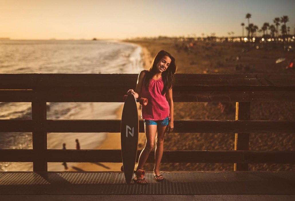 Ivivva Lulu Lemon Girl holding a skateboard standing on a footpath by a beach smiling and posing for the camera looking off sides