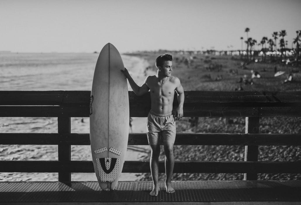 Black and white of young man holding a surfboard on a footpath by a beach posing for a camera and looking offsides