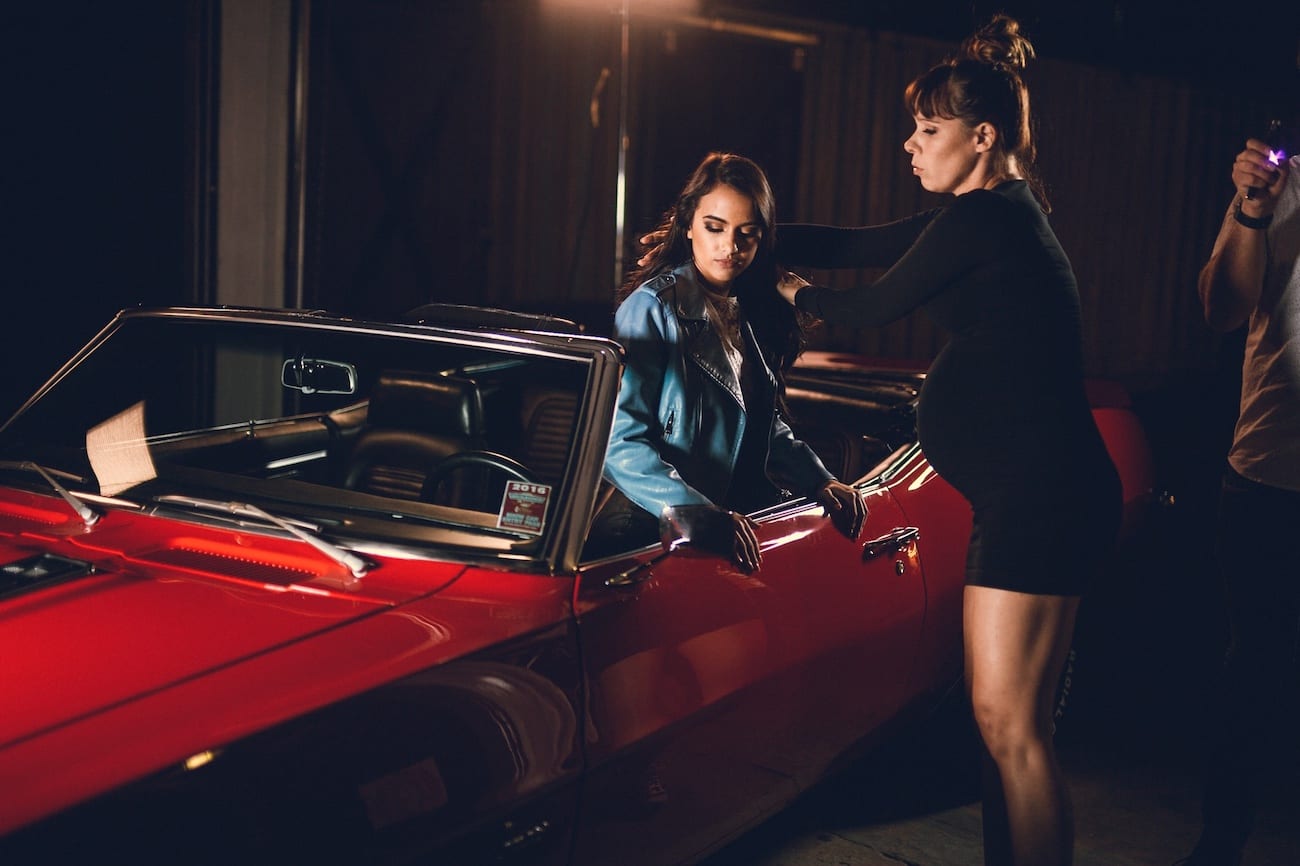 Amy Joy Miller Producer C&I Studios posing for camera kneeling in a vintage car being attended to by a crew member for coat adjustment