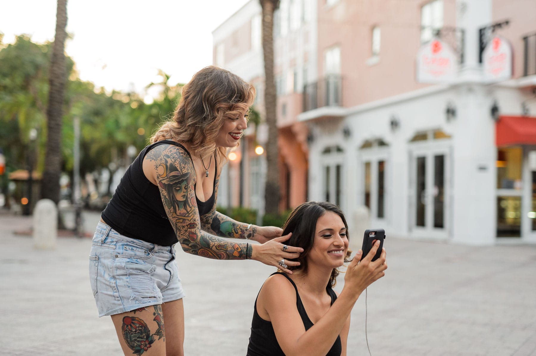 Tattooed makeup artist working on a model who is looking at her cell phone smiling along with the makeup artist
