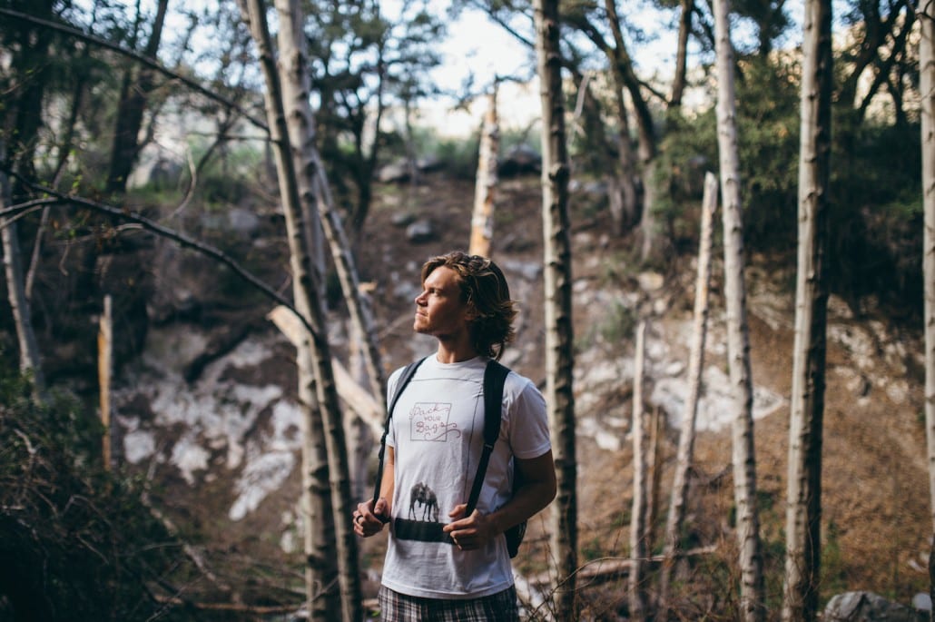 A young man wearing a white tshirt with writing on it and a black backpack among birch trees looking into distance.