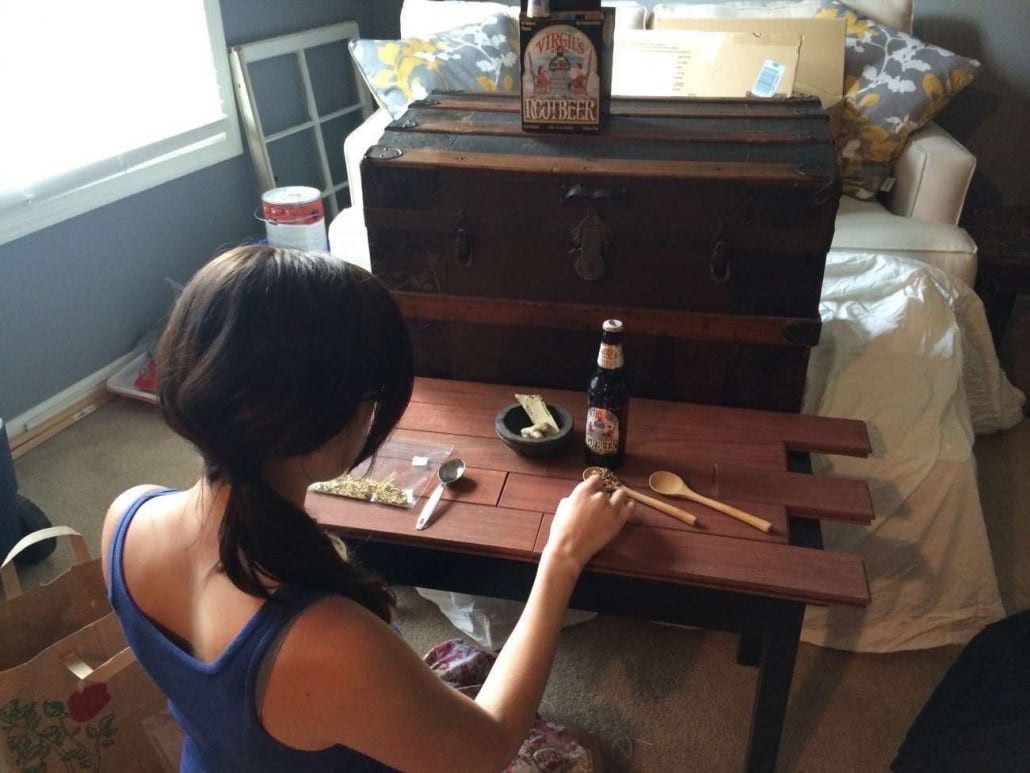 IU CI Studios Portfolio Reeds Bottle of Virgil's Root Beer surrounded by herbs on a wooden table looked upon by a woman kneeling at the table