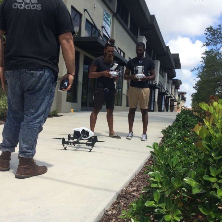 The Manor Two men using drone equipment for a drone on the sidewalk while another man looks on