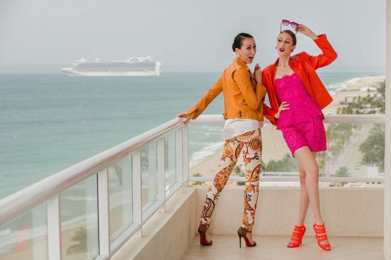 Two women in orange and hot pink outfits posing for camera on a balcony with a cruise ship in the background