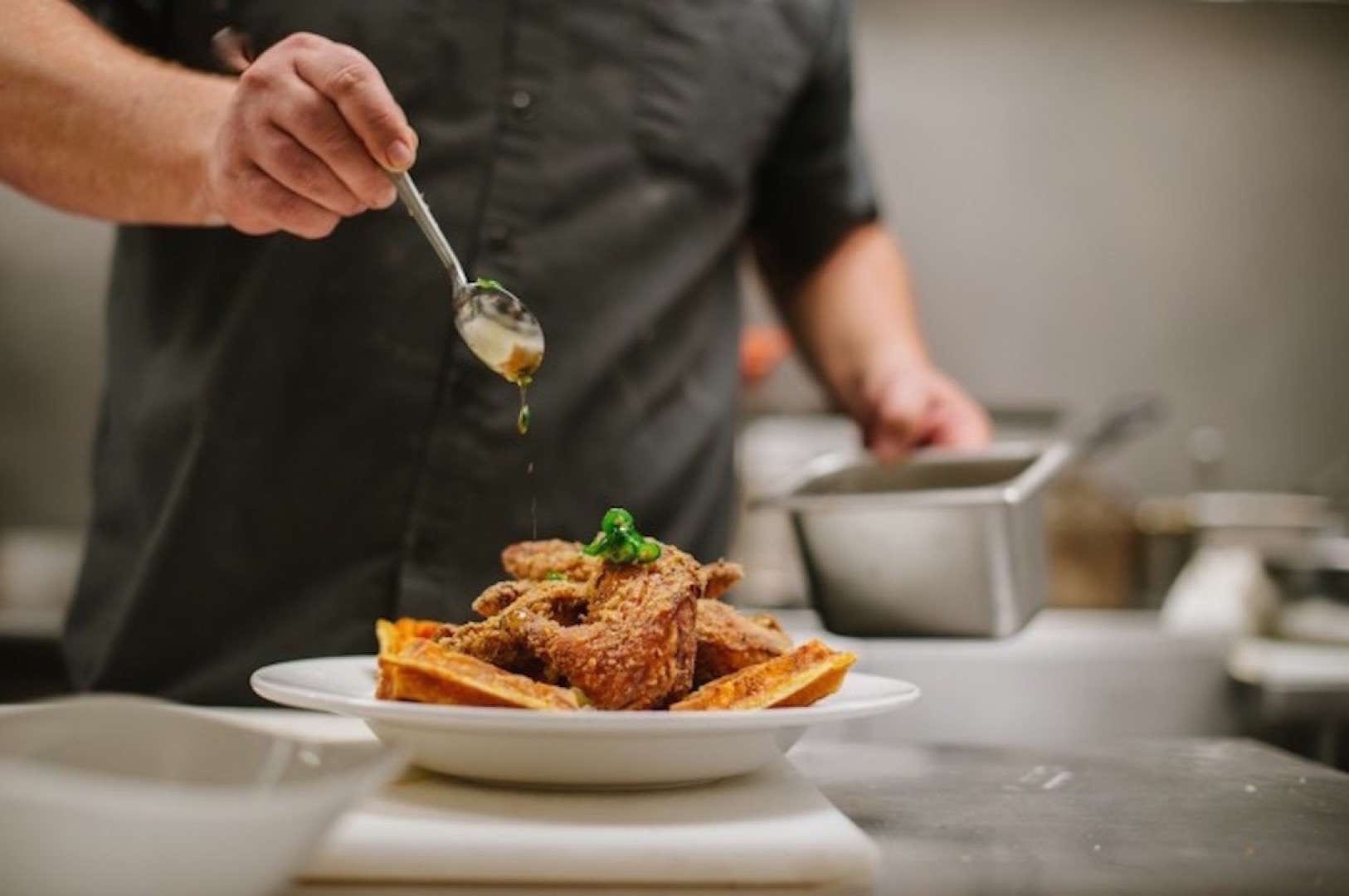 Chef in a black shirt dropping sauce on a plate of fried chicken with potato wedges.