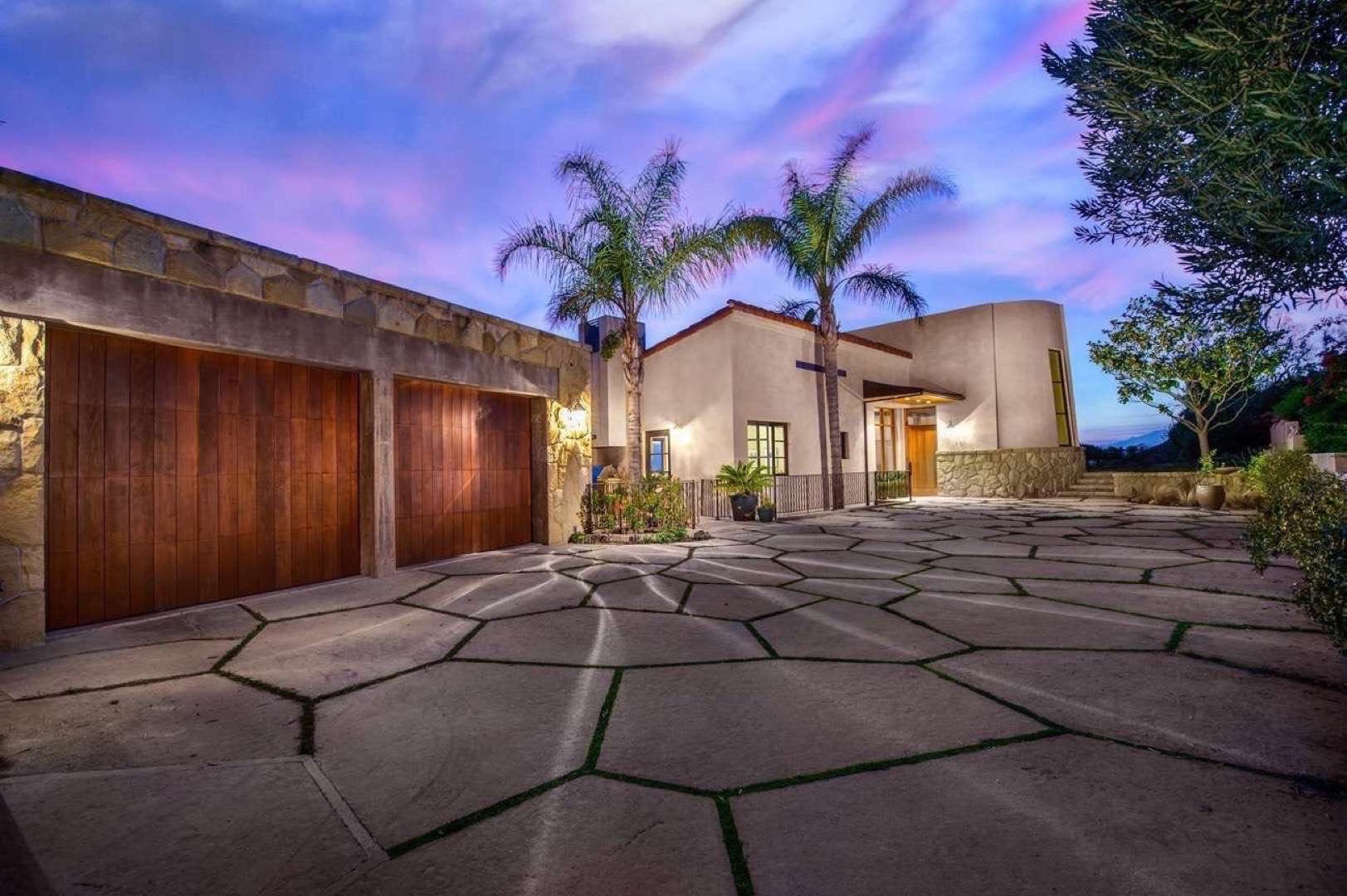 Closeup of home with two car garage with wooden doors and stone patio. There are palm trees and trees around the property.