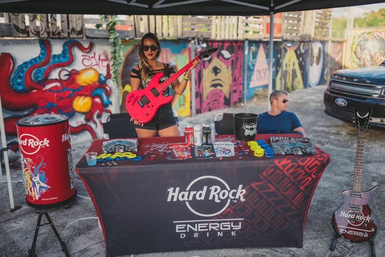 Woman holding red guitar at a Hard Rock Energy Drink booth with a man sitting nearby looking off sides at For the Love Music Festival