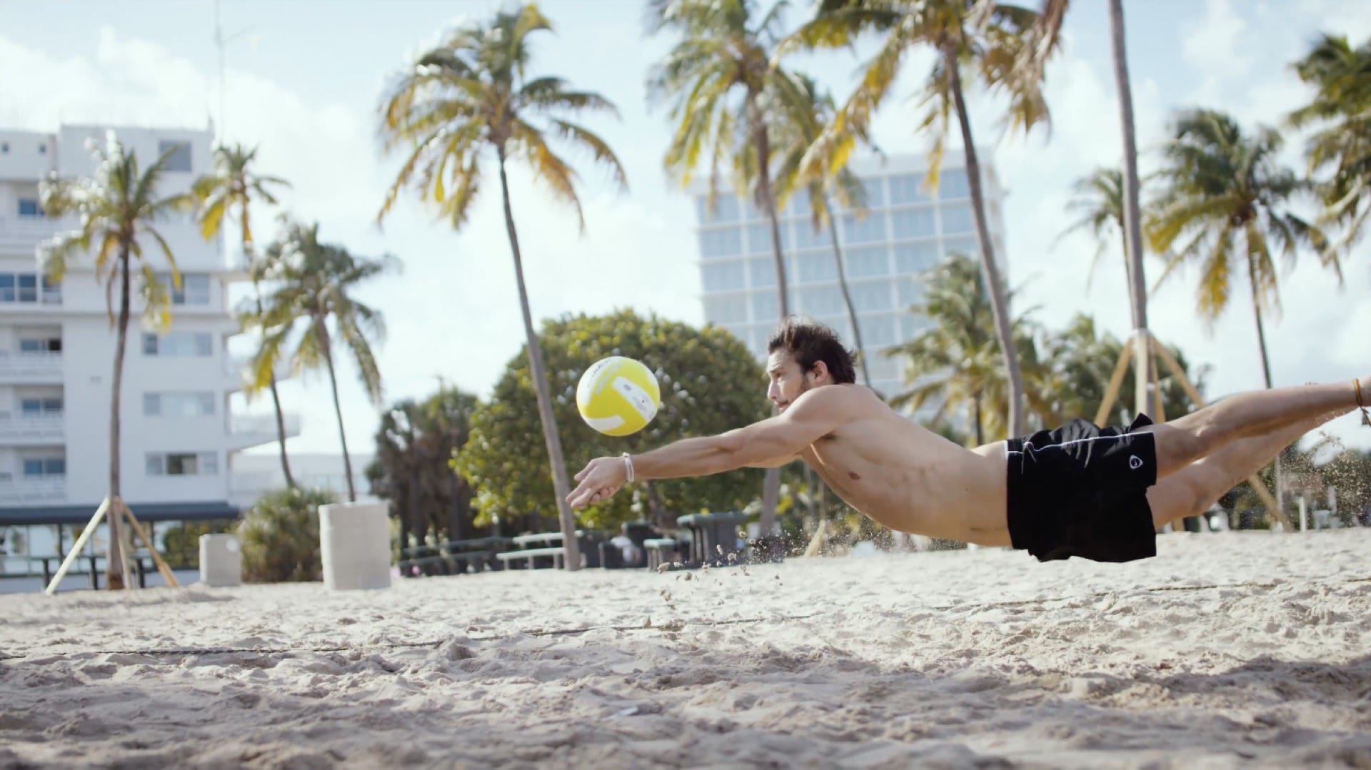 Man with black beach shorts diving to volley a yellow and white volleyball on a beach surrounded by palm trees.