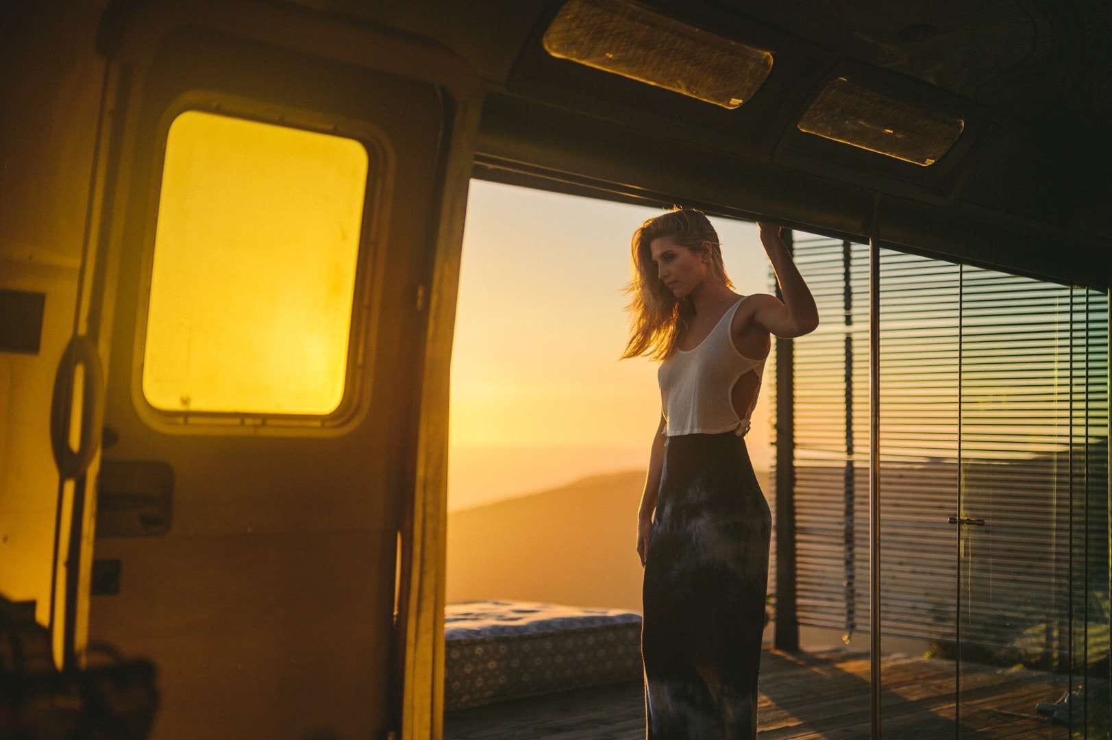 Young woman with long blond hair wearing a white dress shirt and light dress skirt posing from RV at sunrise.