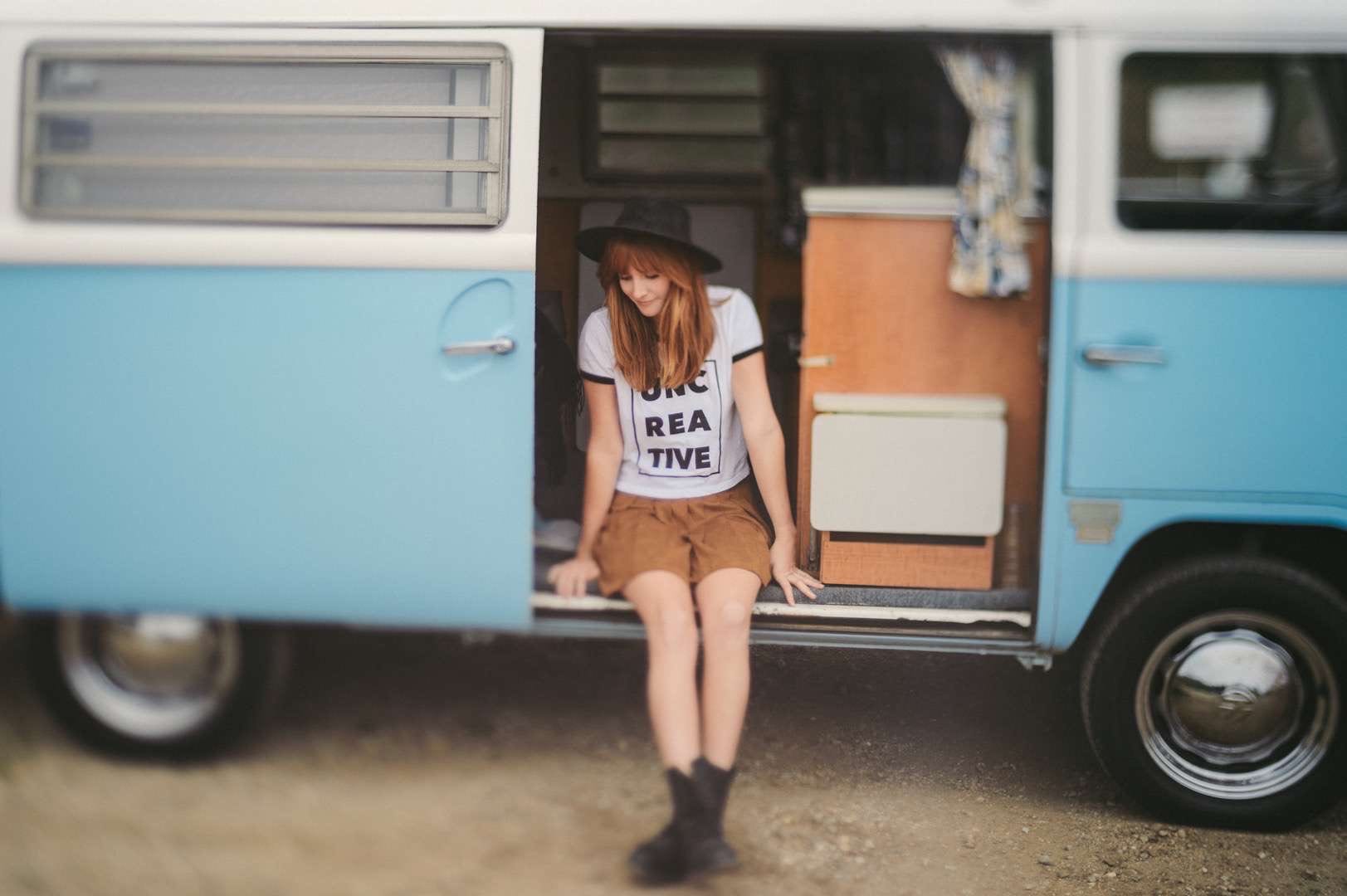 A young woman wearing a white tshirt with Uncreative logo and brown shorts with a black hat looking at the ground. She is sitting in the doorway of a light blue and white RV.