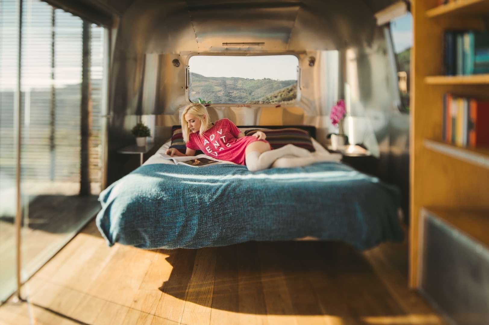 A young woman wearing a red tshirt laying on the bed reading a magazine in a RV.