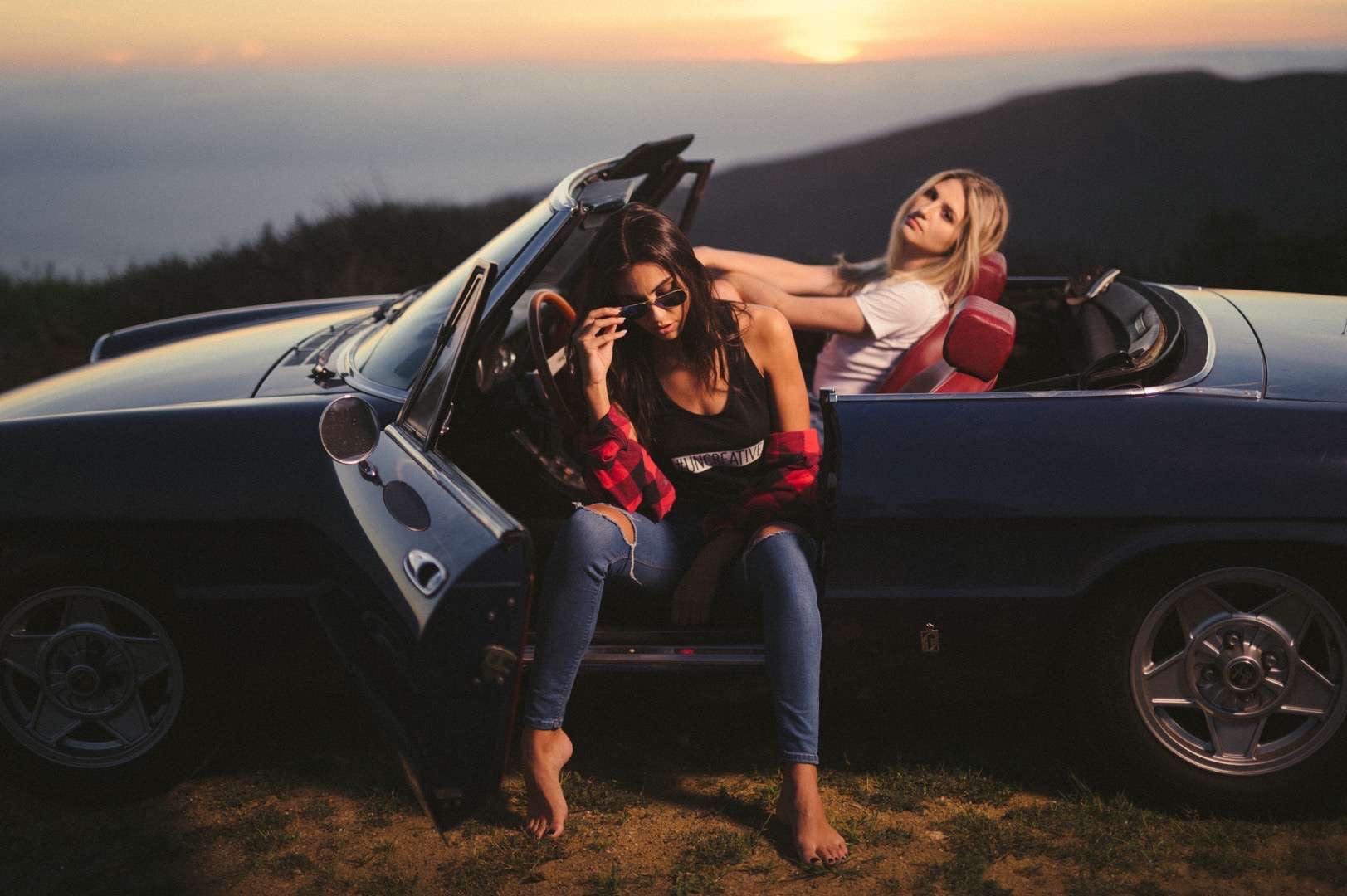 Two young women posing for camera from a dark blue sports car.