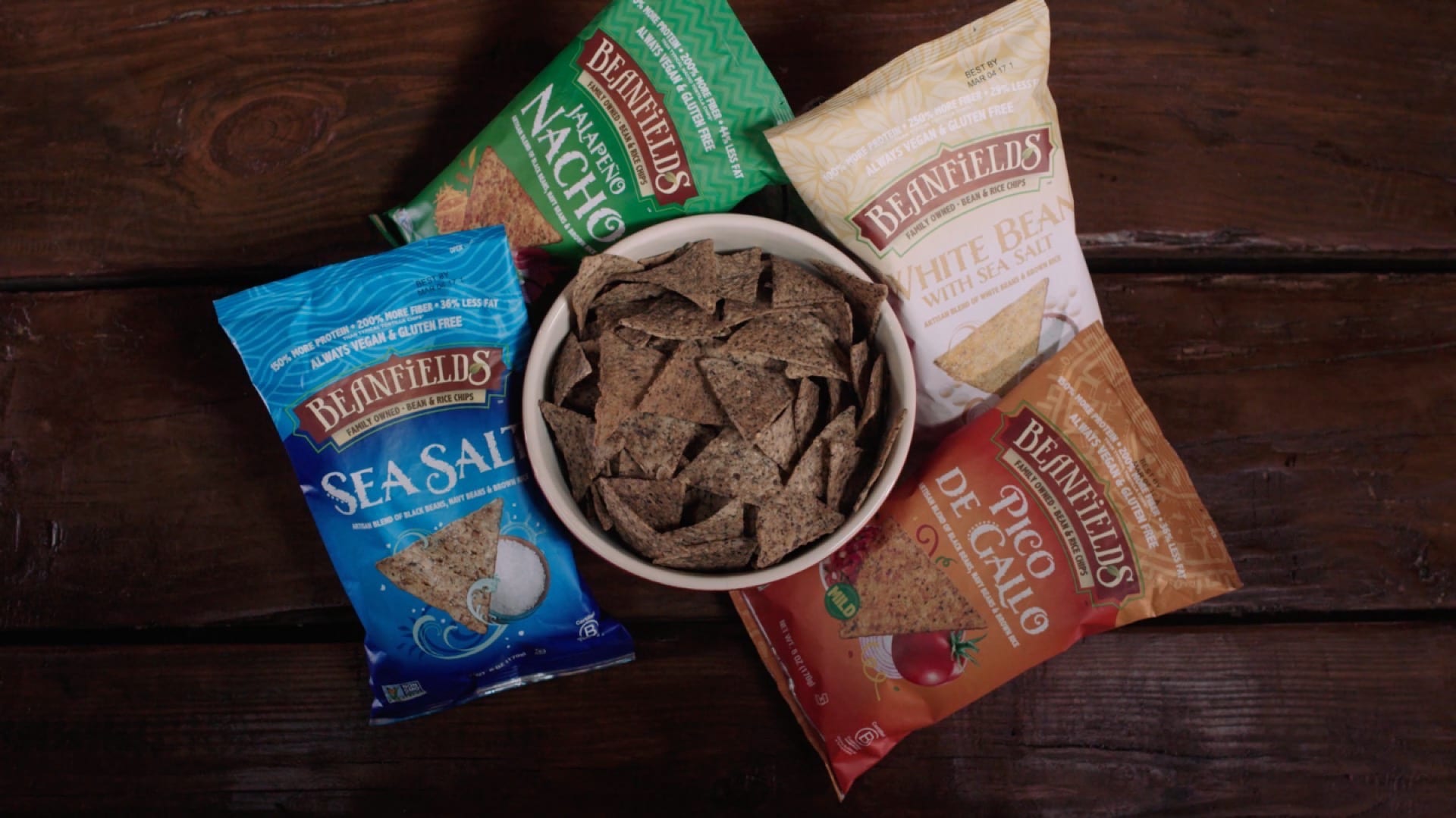 Beanfields Snacks Bowl of brown bean chips surrounded by four bags of Beanfields brand bean chips of different flavors in blue, orange, white and green bags.