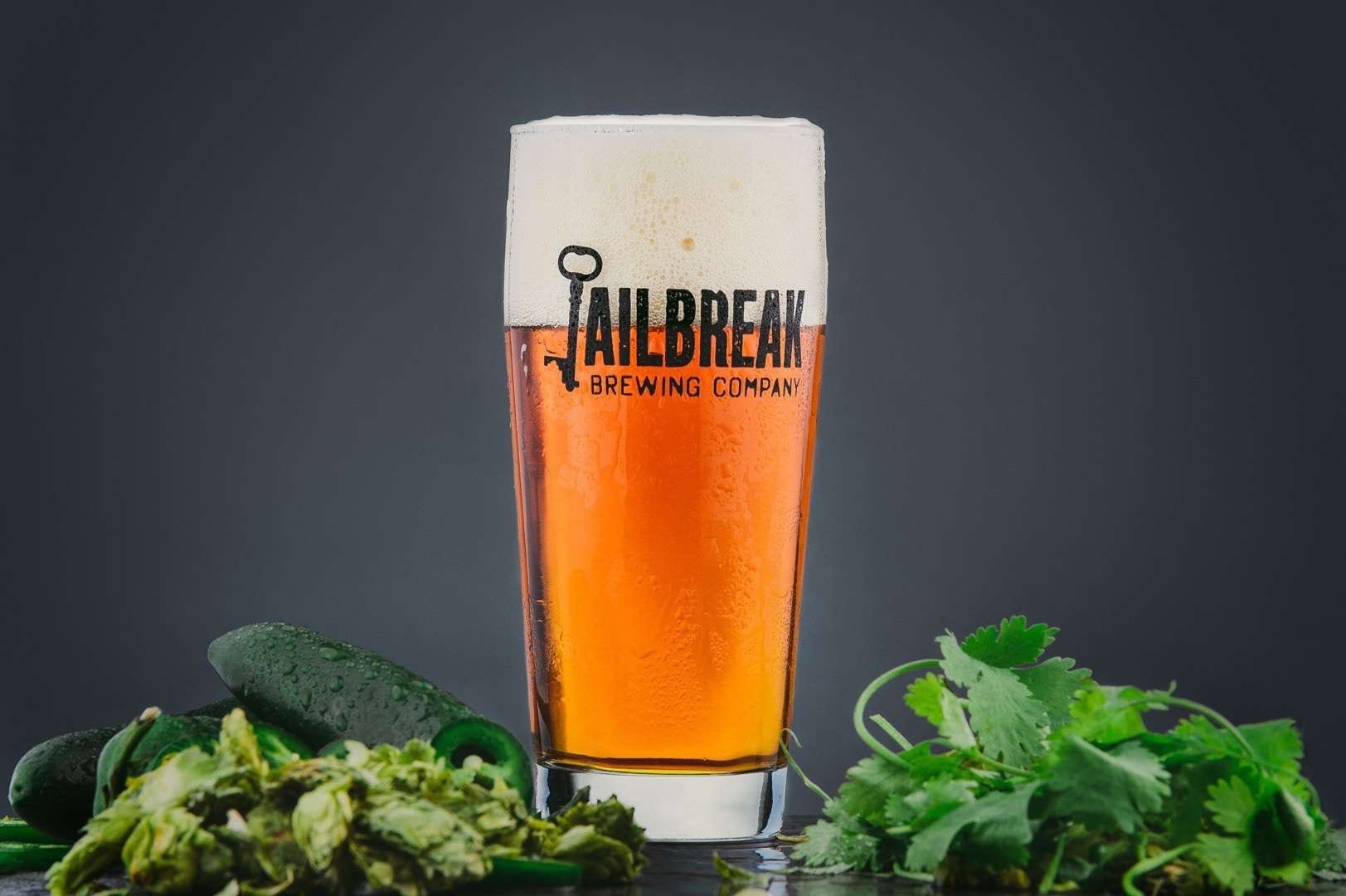 C&I Jailbreak Brewing Closeup of a mostly orange colored glass of ale drink made by Jailbreak Brewing Company. The glass is surrounded by jalapenos and herbs.