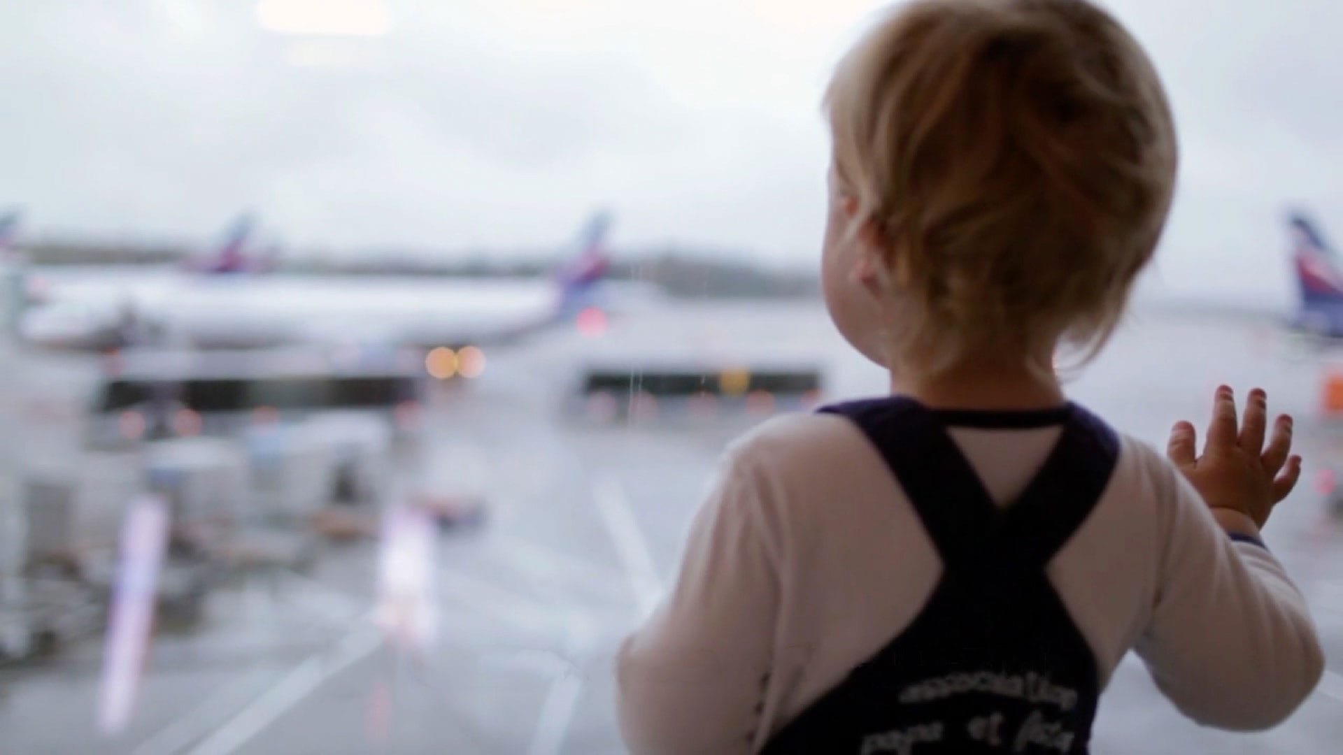 Little boy wearing black coveralls looking out onto the tarmac at a plane at an airport.