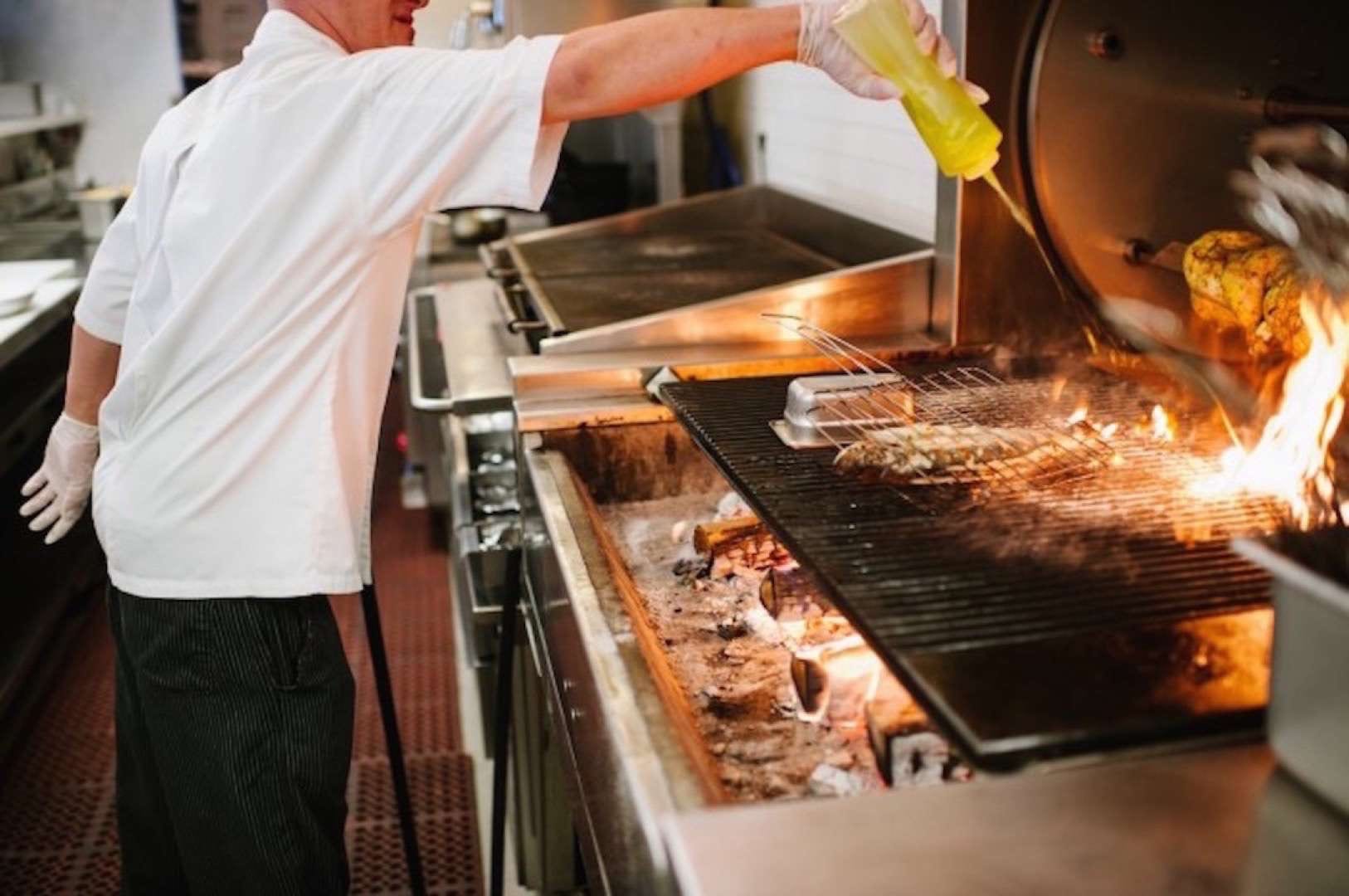 Gulf Stream Media Best New Restaurants Chef wearing latex gloves in a white chef coat and black striped pants spraying oil on a flaming grill.
