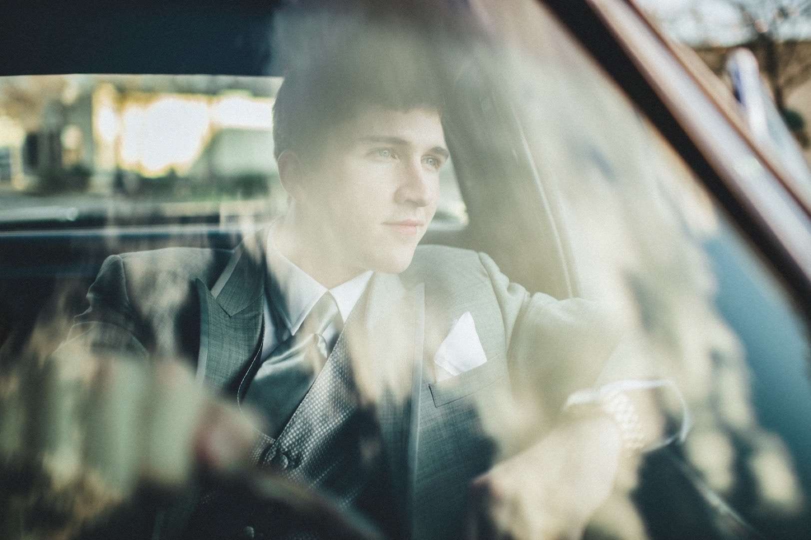 Mens Wearhouse Sprin Lookbook Male model in a gray suit posing for a camera in a car.