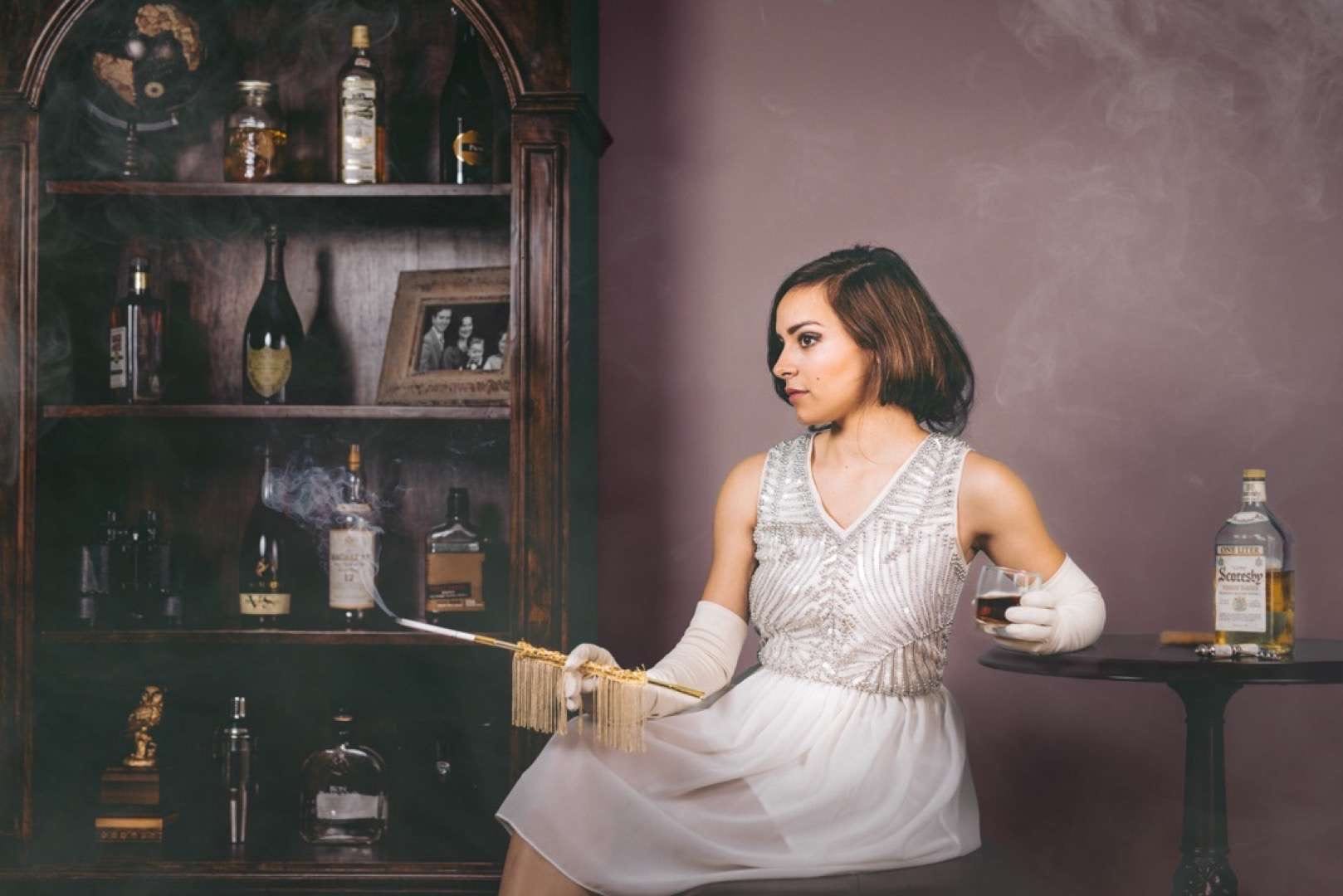 Woman in an old setting with a vintage cigarette posing for the camera with a glass of alcohol in her hand. There is a bottle of alcohol on the table nearby and a shelf with alcohol bottles and other items.