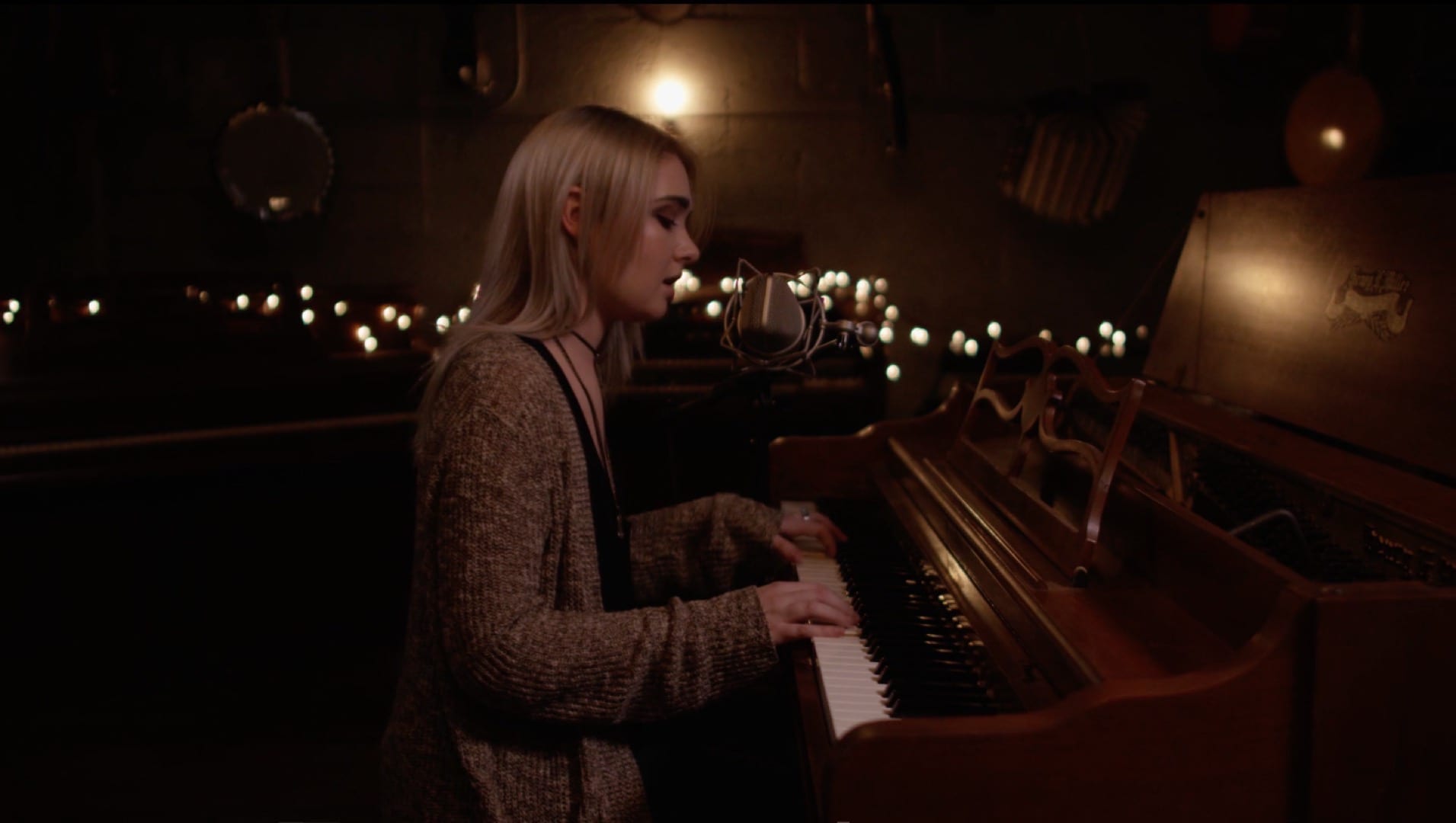 Woman with long blond hair and wearing a beige knitted coat playing a piano and singing in a candlelight scene.