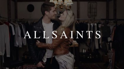 CI Studios Portfolio White All Saints Logo On Dimmed Background Of Young Man And Woman Looking At Each Other In The Eyes Smiling Embracing Each Other Surrounded By Clothes Racks With Clothes