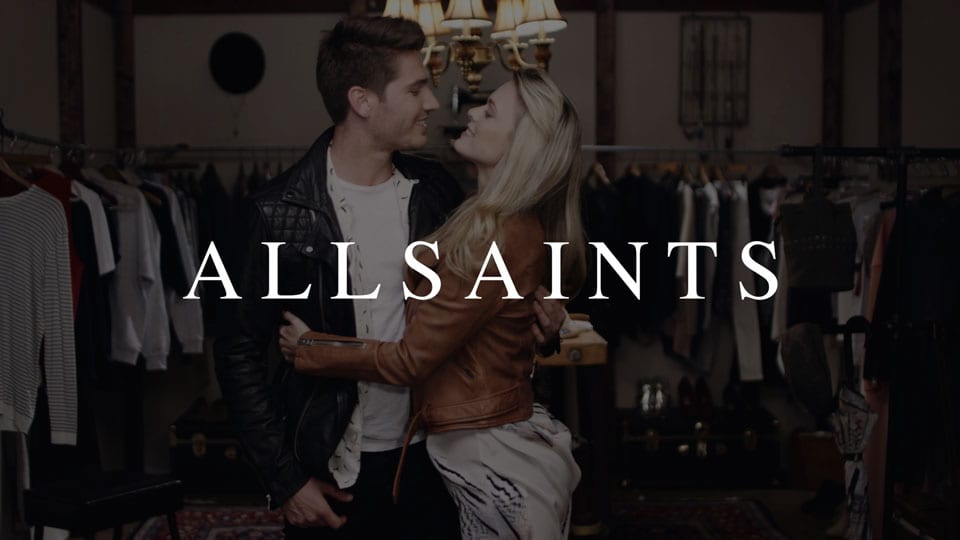 CI Studios portfolio White All Saints logo on dimmed background of young man and woman looking at each other in the eyes smiling embracing each other surrounded by clothes racks with clothes