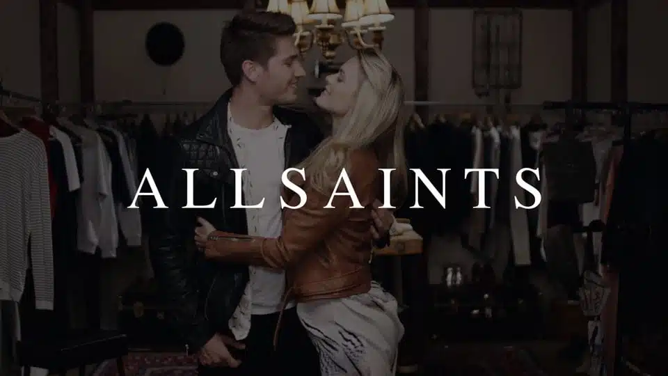 IU C&I Studios Page CI Studios portfolio White All Saints logo on dimmed background of young man and woman looking at each other in the eyes smiling embracing each other surrounded by clothes racks with clothes
