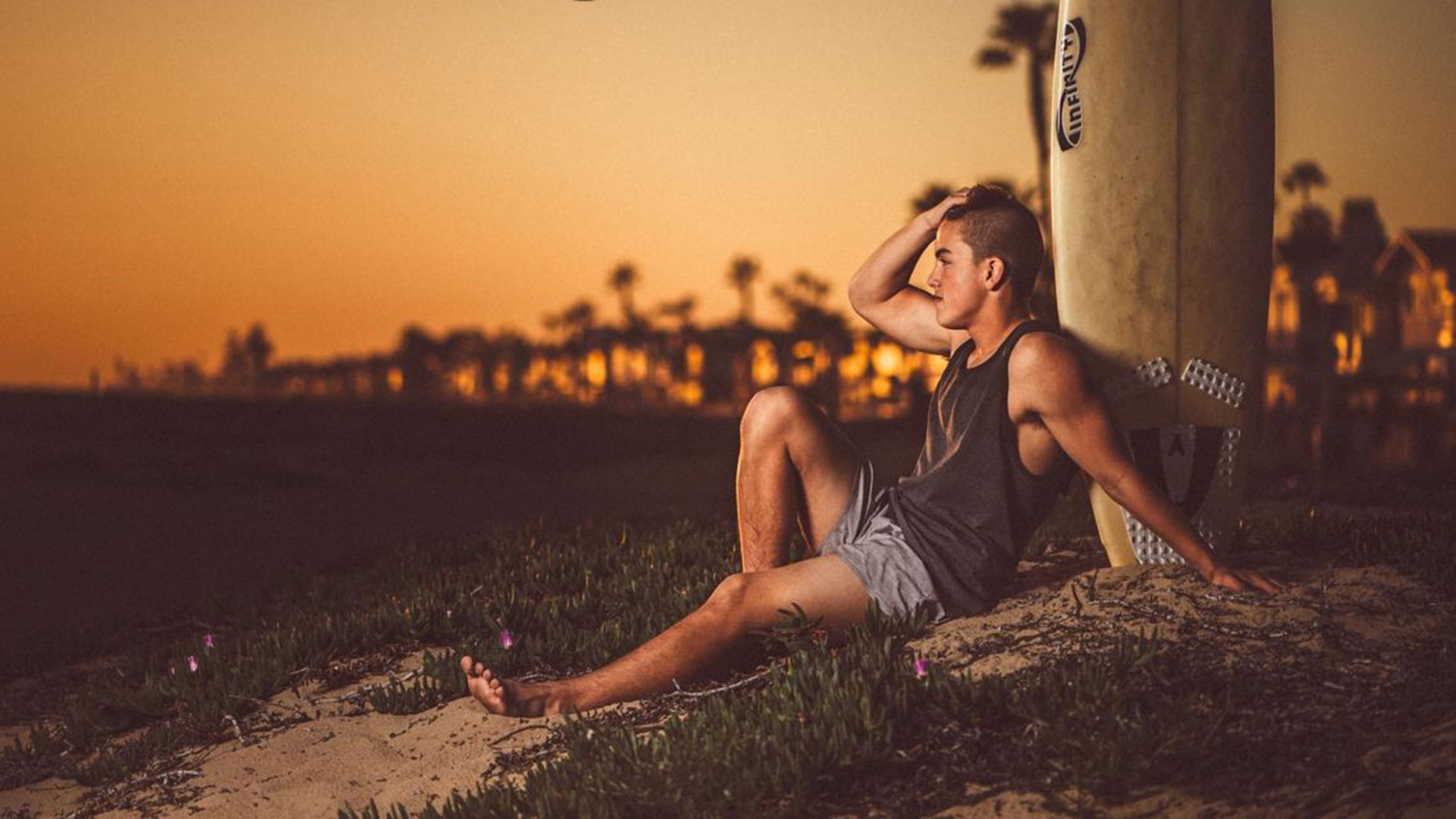 Young guy wearing a black tank top and gray shorts leaning against a surfboard looking off to the distance at dusk