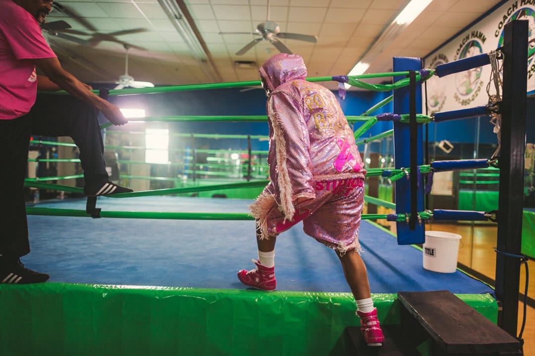 Woman in pink outfit climbing onto the blue boxing mat with light green boundaries. Her training is climbing in nearby. There is a white bucket on the mat nearby.