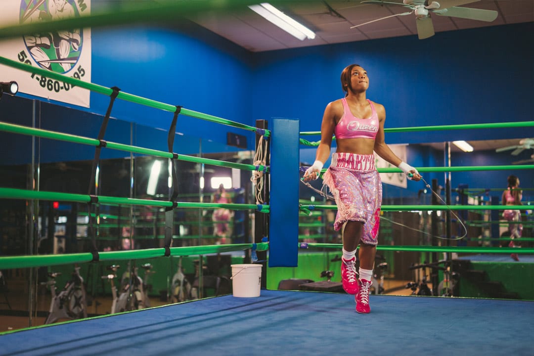 Gold Coast Boxer Female fighter in pink outfit jumping rope dark blue mat with light green boundaries. She is wearing bright red shoes. There are some exercise bicycles and mirrors in the background.