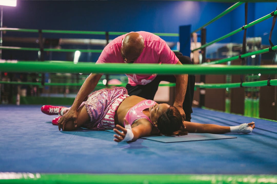 Female fighter in pink outfit lying down on dark blue mat with light green boundaries. She is wearing bright red shoes. Her trainer is helping with leg related stretching.