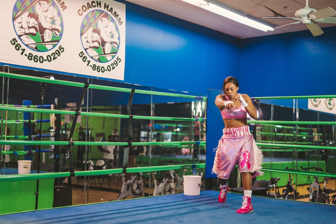 Female fighter in pink outfit practicing boxing moves on a dark blue mat with light green boundaries. She is wearing bright red shoes. There are some exercise bicycles and mirrors and a white bucket in the background.