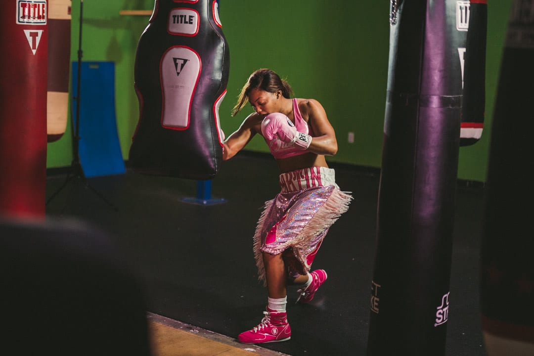 Gold Coast Boxer Woman in pink outfit with pink boxing gloves training with a semi-large punching bag in a gym with a few big punching bags.