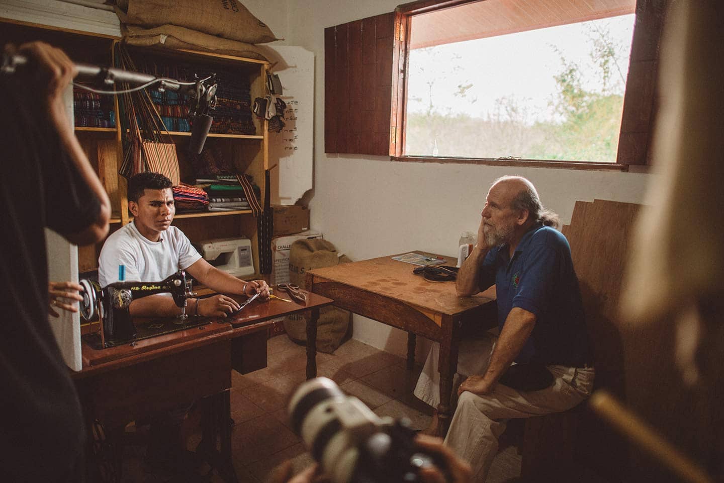 IU C&I Studios Page Man using a sewing machine in a small tailor shop building by a window with sun streaming in. There is another man sitting nearby and a film crew filming.