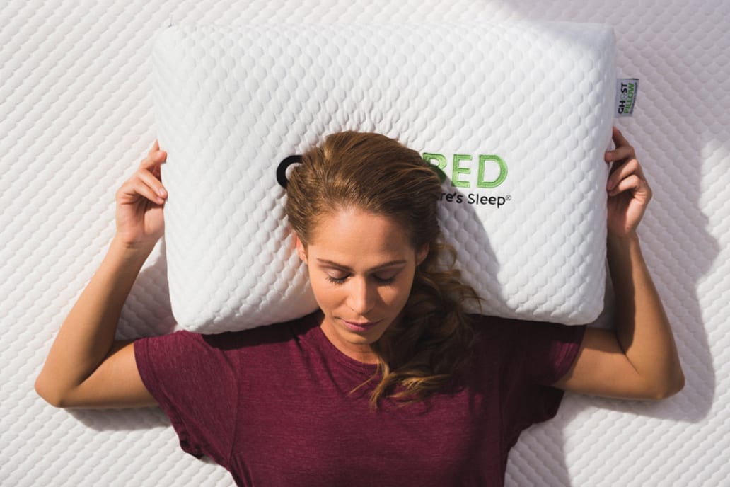 Ghostbed Woman posing for camera with a Ghostbed pillow on a bed lying on it with eyes closed and arms outstretched around it