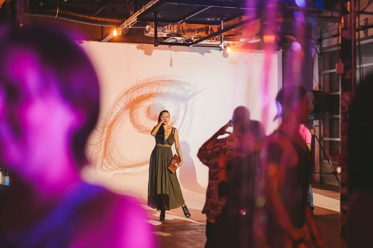C&I Storm C&I Storm at FATVillage Artwalk Asian woman in olive dress posing for camera in front of backdrop that has a drawing of an eye on it