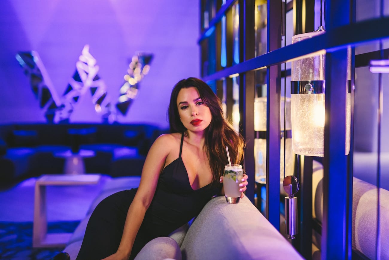 Woman with long brown hair wearing a black dress sitting by the bat with a drink posing for the camera