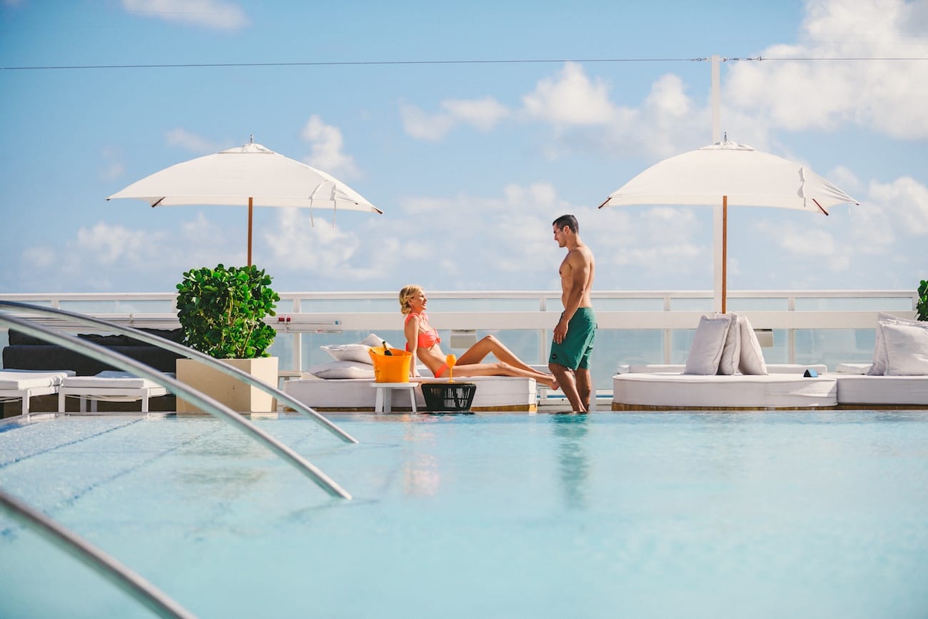 Man in green swimming trunks talking to a woman in pink bikini sitting on a lounge chair