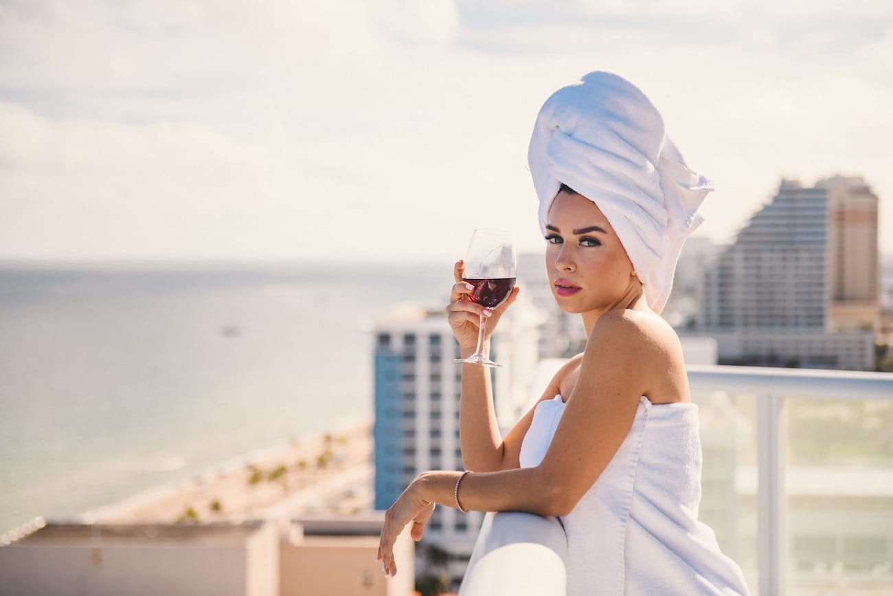 Woman wearing white towels on body and head enjoying a glass of wine leaning on a balcony railing posing for the camera