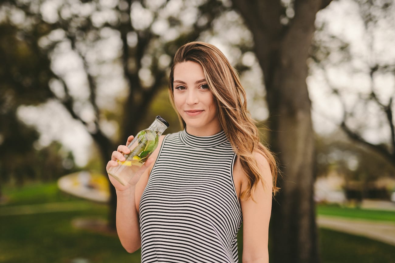 Juicery RX Marketing Solutions by C&I An Idea Agency Woman with long blond hair in a black and white striped tank top holding a glass bottle of water with herbs in it smiling and posing for the camera