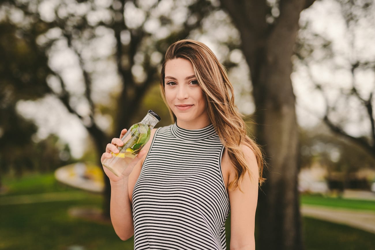 Woman with long blond hair in a black and white striped tank top holding a glass bottle of water with herbs in it smiling and posing for the camera